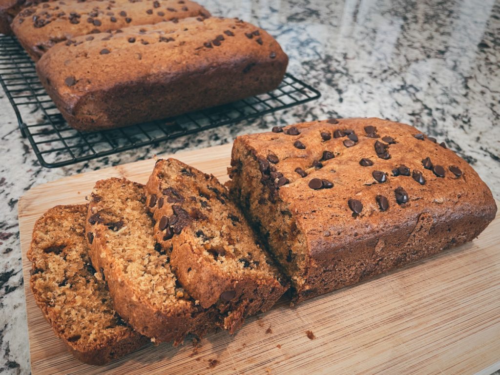a half-sliced loaf of banana bread sits on a cutting board in the foreground with 2 more loaves on a cooling rack in the background
