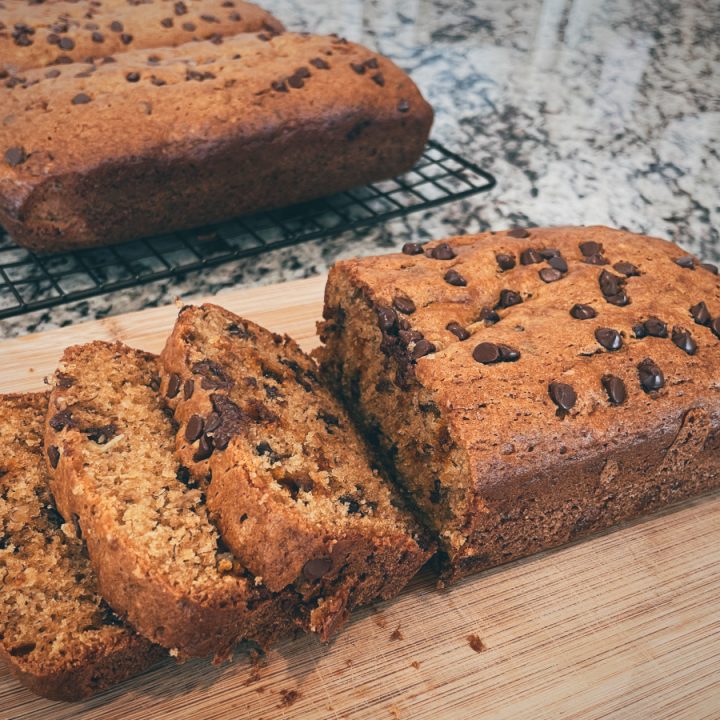 a half-sliced loaf of banana bread sits on a cutting board in the foreground with 2 more loaves on a cooling rack in the background