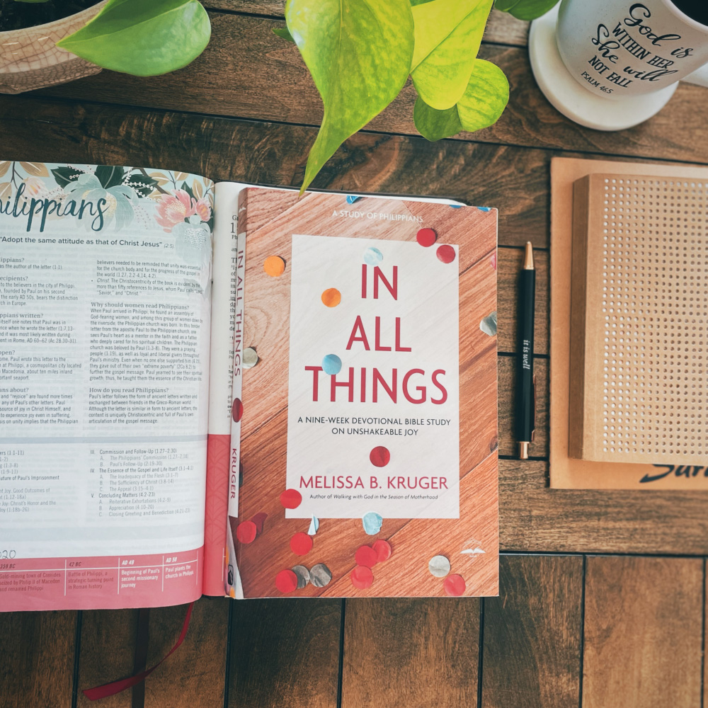 In All Things by Melissa B. Kruger book on a table with a Bible open to the book of Philippians with a journal, a mug of coffee, and a green pothos plant on the table next to it.