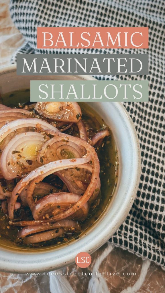 close up of sliced shallots in a bowl with olive oil, herbs, and spices in a white bowl sitting on top of a wooden cutting board with black and white cloth napkin aside with words "Balsamic Marinated Shallots" overlaid