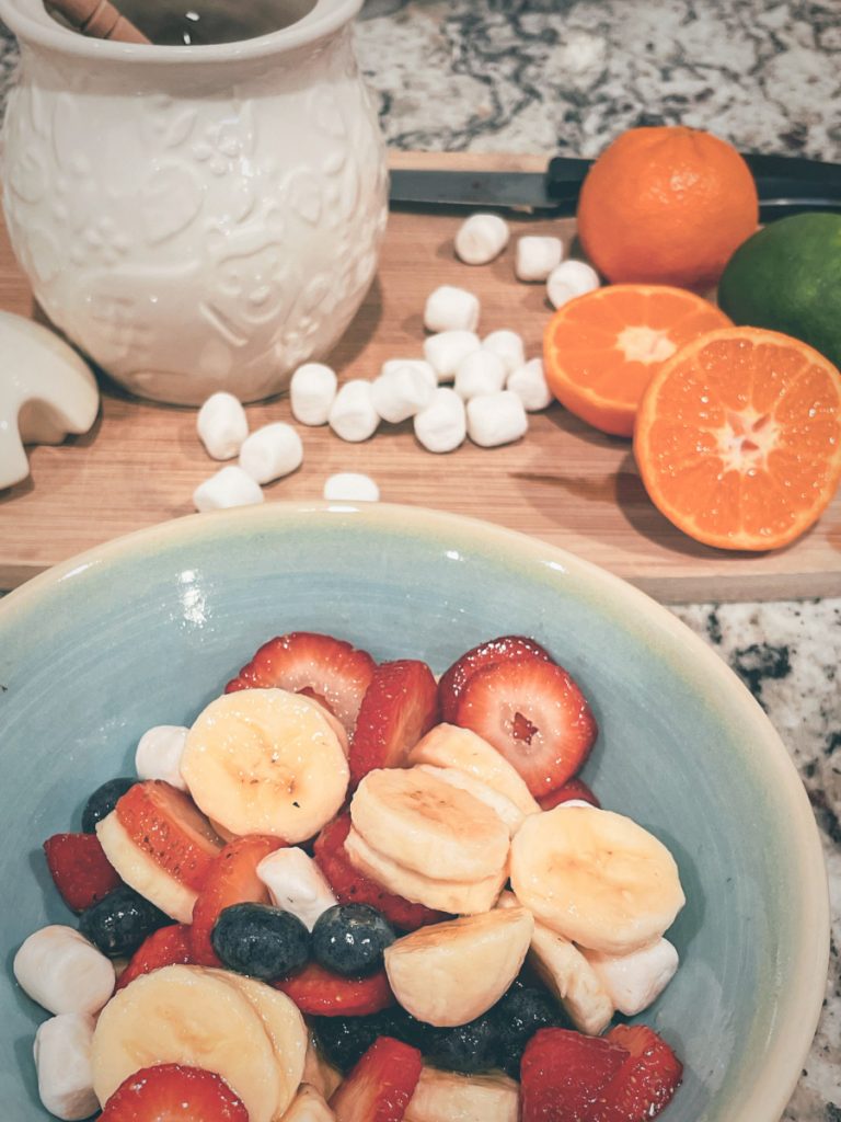 simple summer fruit salad consisting of bananas, strawberries, blueberries, and marshmallows in a blue bowl next to a cutting board with a honey pot, marshmallows, sliced oranges and limes