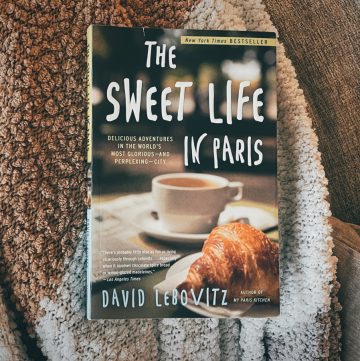 "The Sweet Life in Paris" book laying on a nubby gray, white, and tan blanket on a couch