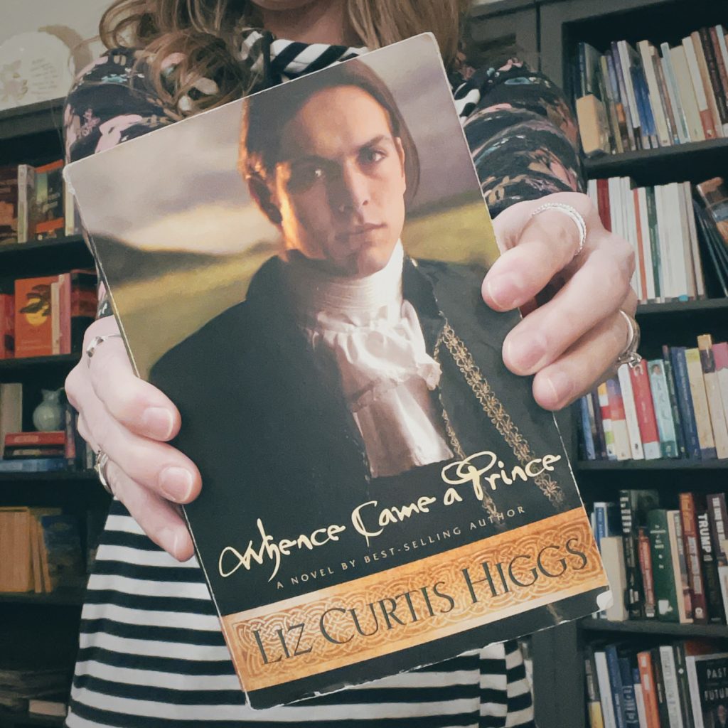 woman standing in front of a bookshelf, holding out the book "Whence Came a Prince" by Liz Curtis Higgs