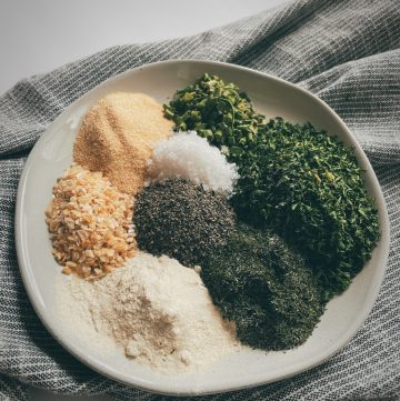 a small cream colored plate on top of a grey and white cloth napkin with dried herbs and seasonings that are the ingredients to make homemade ranch seasoning in small piles