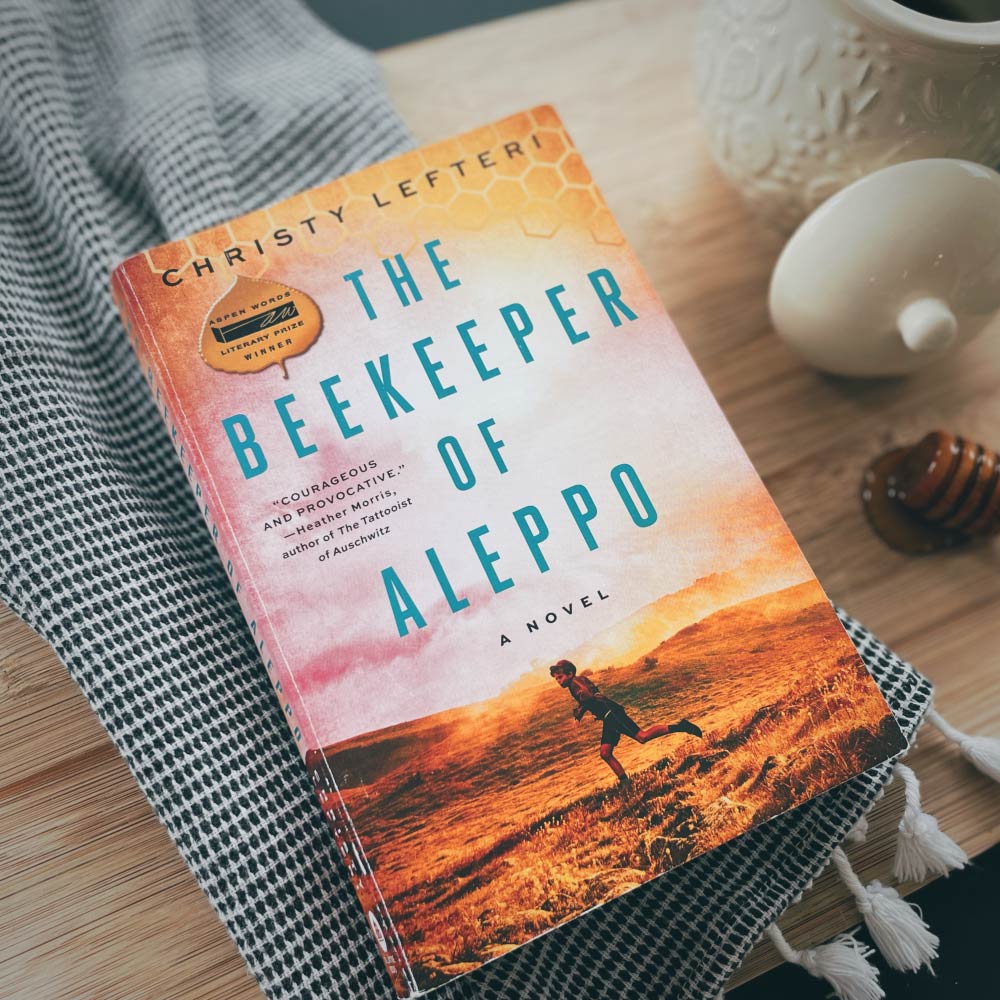 photo of the cover of the Beekeeper of Aleppo book on top of a black and white cloth next to a honey jar