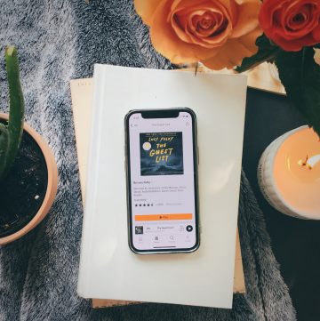 image of a phone with the audible app open showing the front cover of The Guest List by Lucy Foley sitting on a stack of books with a plant, candle, and flowers around it