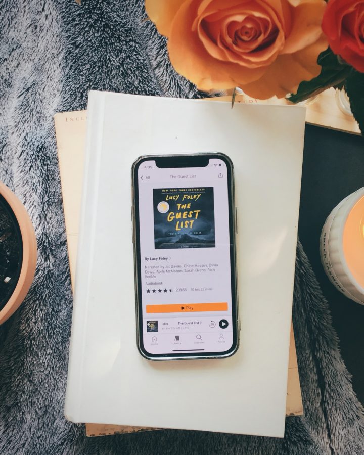 image of a phone with the audible app open showing the front cover of The Guest List by Lucy Foley sitting on a stack of books with a plant, candle, and flowers around it