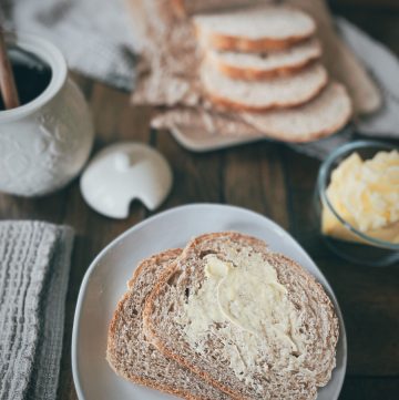 close up of two slices of whole wheat sourdough bread with butter on a cream colored round plate in the foreground with a honey pot, butter dish, and a sliced loaf of bread in the background on a wooden table