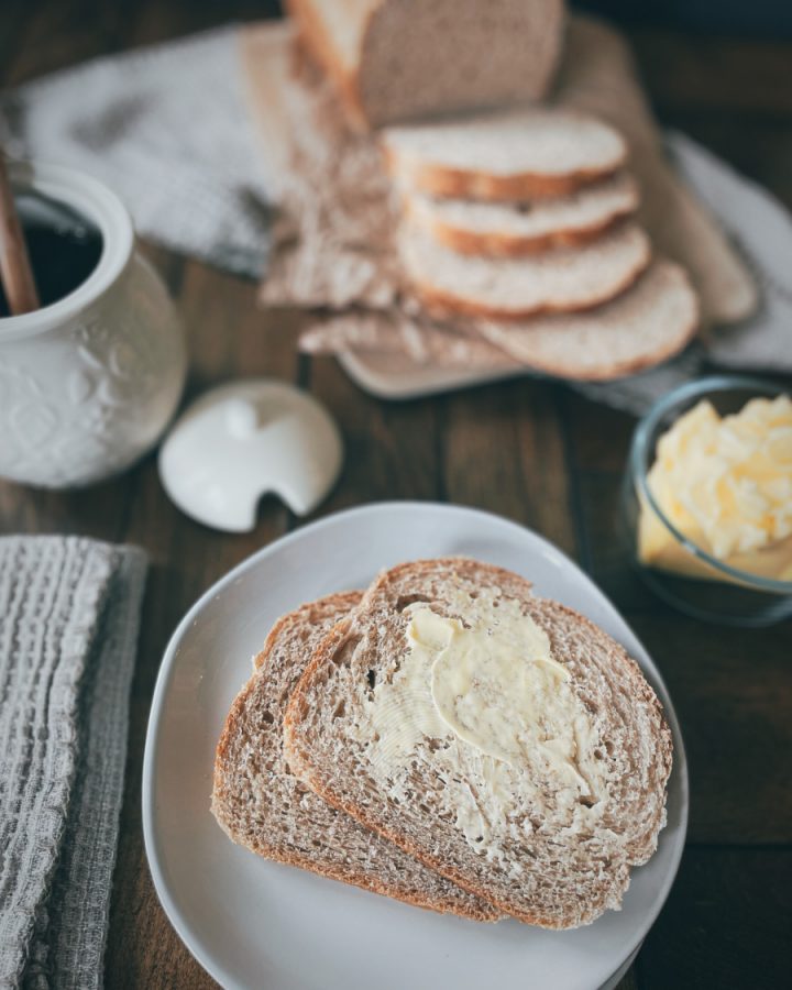 close up of two slices of whole wheat sourdough bread with butter on a cream colored round plate in the foreground with a honey pot, butter dish, and a sliced loaf of bread in the background on a wooden table