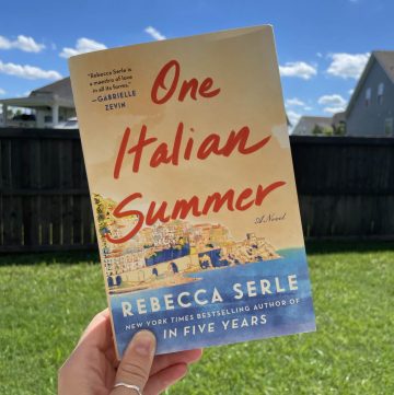 image of a hand holding the book One Italian Summer with green grass and a blue sky in the background