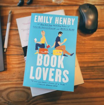 photo of the cover of Book Lovers by Emily Henry on top of a wooden desk next to a computer, pen, and mouse
