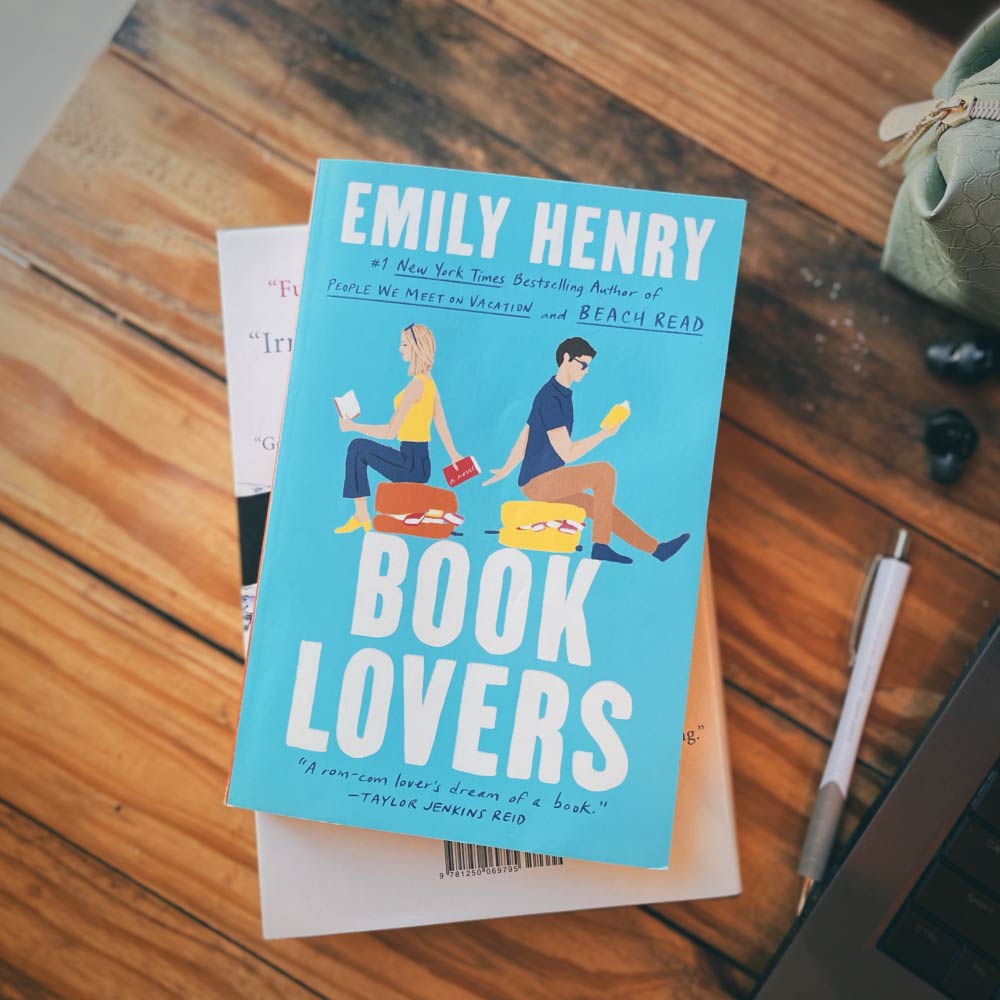 photo of the cover of Book Lovers by Emily Henry on top of a wooden desk next to a pen and pencil case