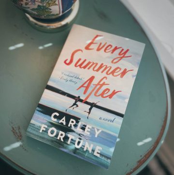 photo of the cover of Every Summer After by Carley Fortune sitting on a teal side table next to a colorful lamp